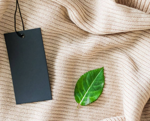 Ausschnitt eines hellbraunen Kleidungsstücks, darauf ein Preislabel und ein grüne Blatt – Symbolbild für nachhaltige Preisstrategie im E-Commerce.
