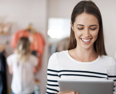Zeitmanagement im E-Commerce: Zufriedene Onlinehändlerin schaut glücklich auf Ihr Tablet. Im Hintergrund weitere Frau, die sich im Shop für Kleidung um Bekleidung am Bügel kümmert.