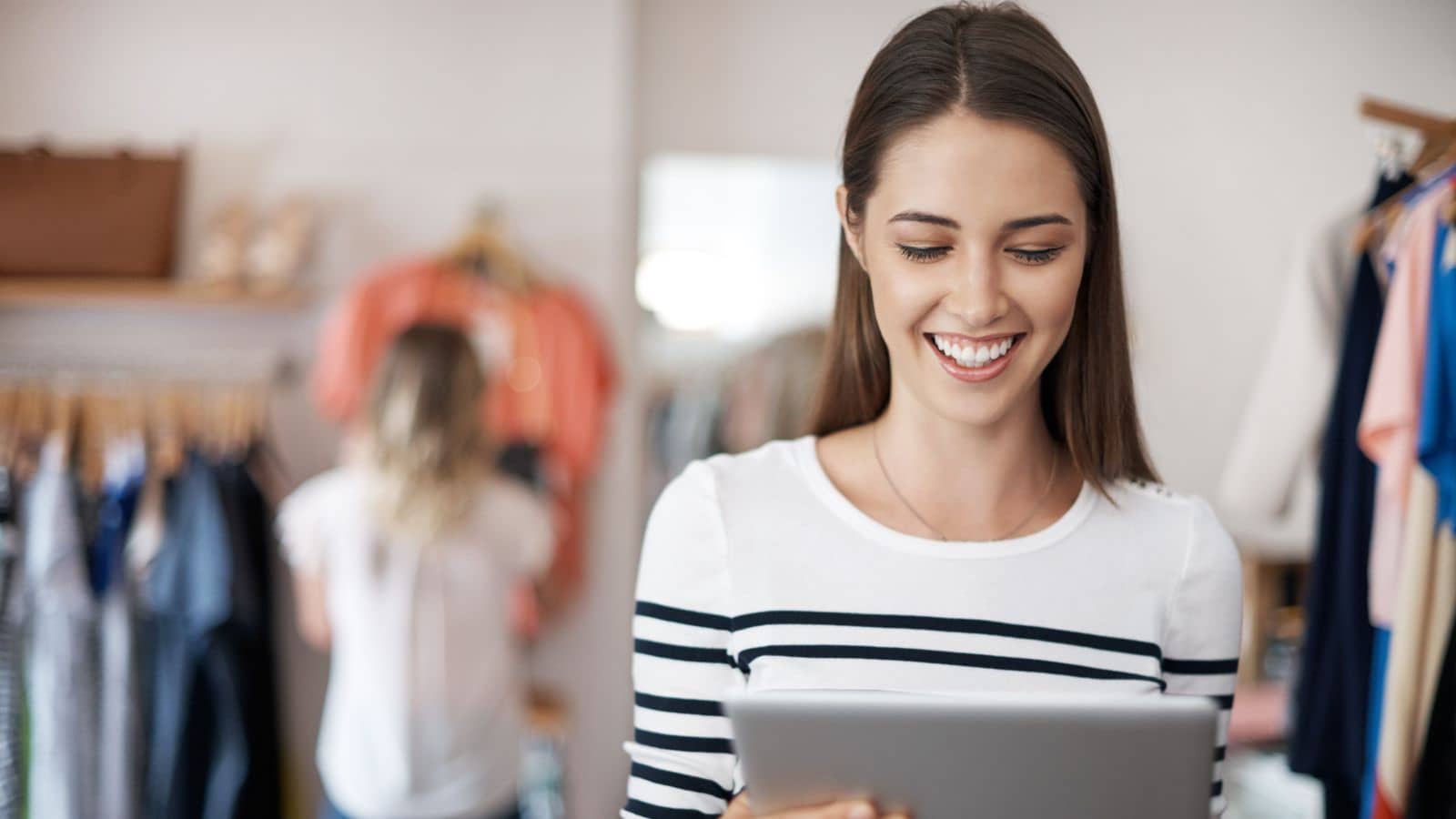 Zeitmanagement im E-Commerce: Zufriedene Onlinehändlerin schaut glücklich auf Ihr Tablet. Im Hintergrund weitere Frau, die sich im Shop für Kleidung um Bekleidung am Bügel kümmert.
